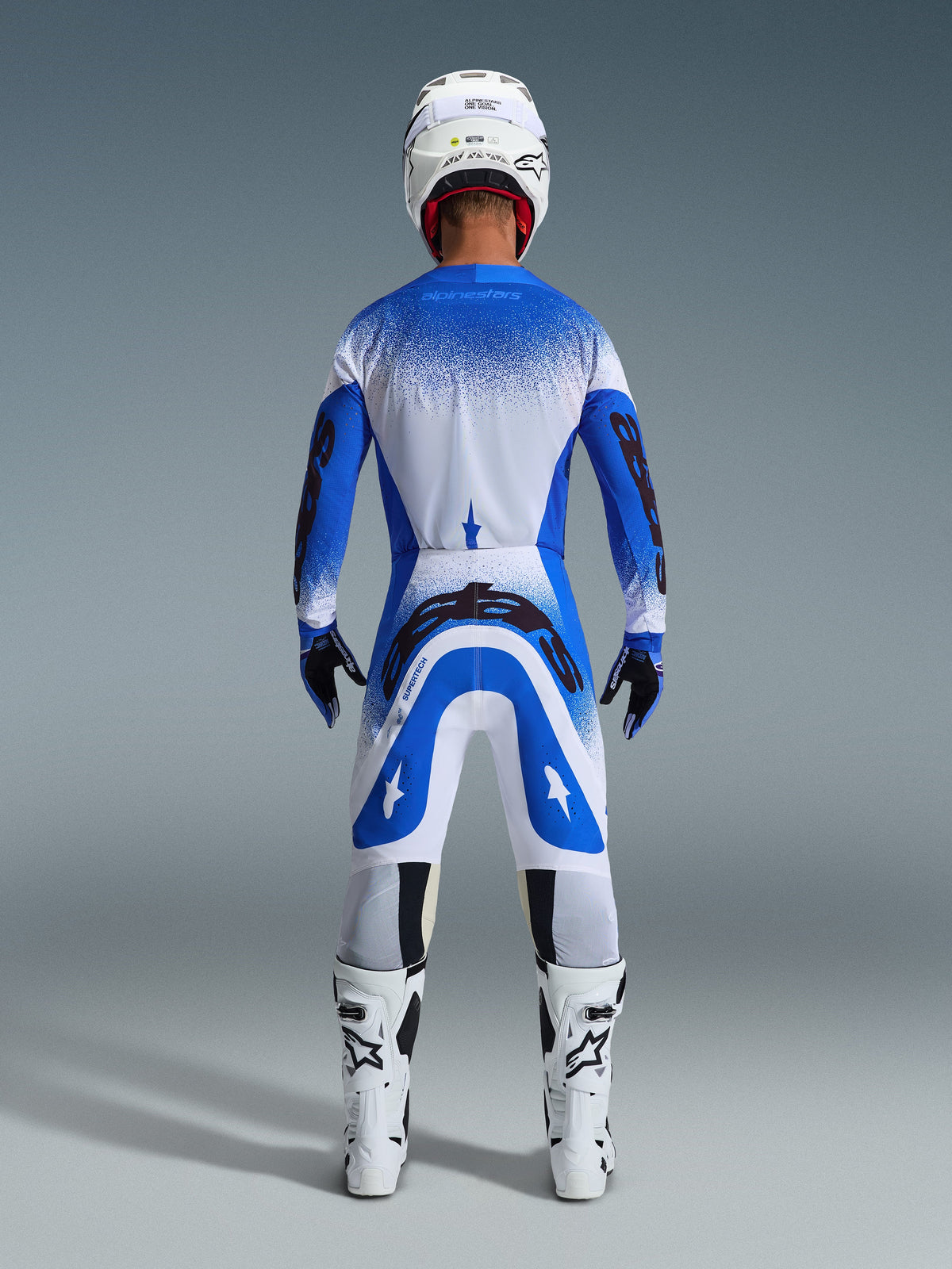 Motocross rider from the back wearing the Alpinestars 2026 Supertech Scenz Gear Set in Ucla Blue White, blue and white jersey with black branding, matching pants, white helmet, and white boots, coordinated pro-level racing gear for off-road motocross