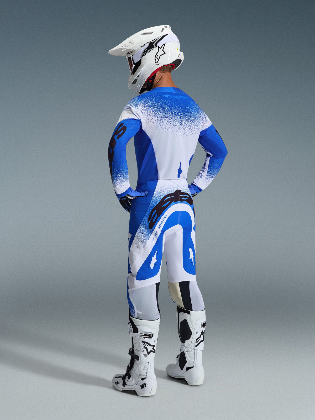 Alpinestars 2026 Supertech Scenz Gear Set, back view of a rider wearing the Ucla Blue White jersey and pants, white motocross helmet, and white racing boots, pro-level racing performance outfit