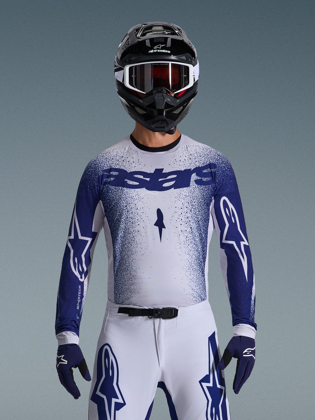 Motocross rider wearing the Alpinestars 2026 Supertech Scenz Gear Set, white jersey with a blue speckled gradient and 'astars' branding, matching white pants with blue accents, black helmet with white goggles, and blue gloves, showing the athletic pro-level racing fit.
