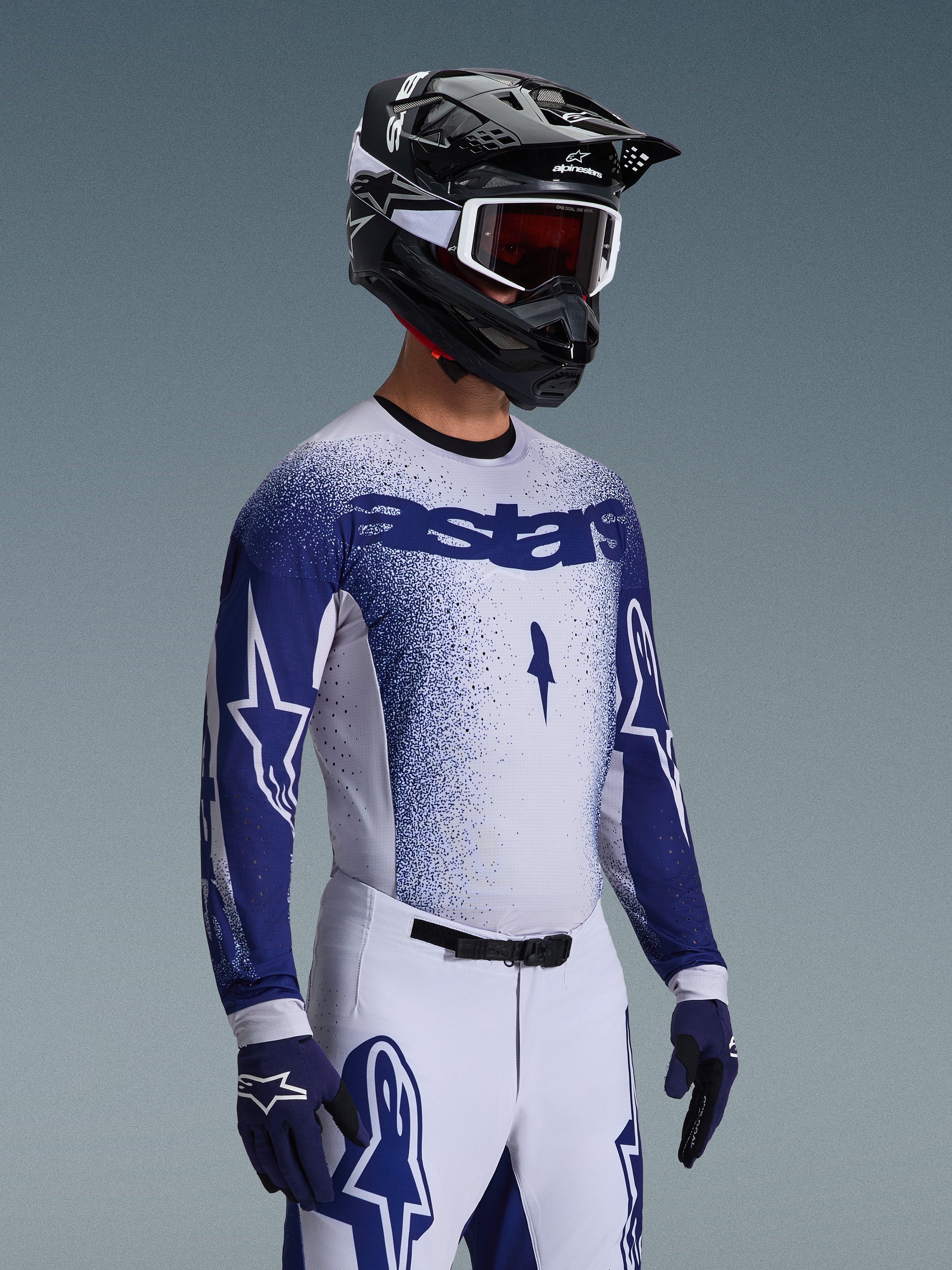Motorcrossrijder met de 2026 Supertech Scenz Jersey in lichtgrijs en donkerblauw, gecombineerd met een bijpassende witte en blauwe racebroek, blauwe handschoenen en een zwarte helm met witte crossbril. De jersey heeft een gestippeld blauw verloop-effect op de borst en mouwen, een professionele atletische pasvorm en opvallende 'astars' branding.