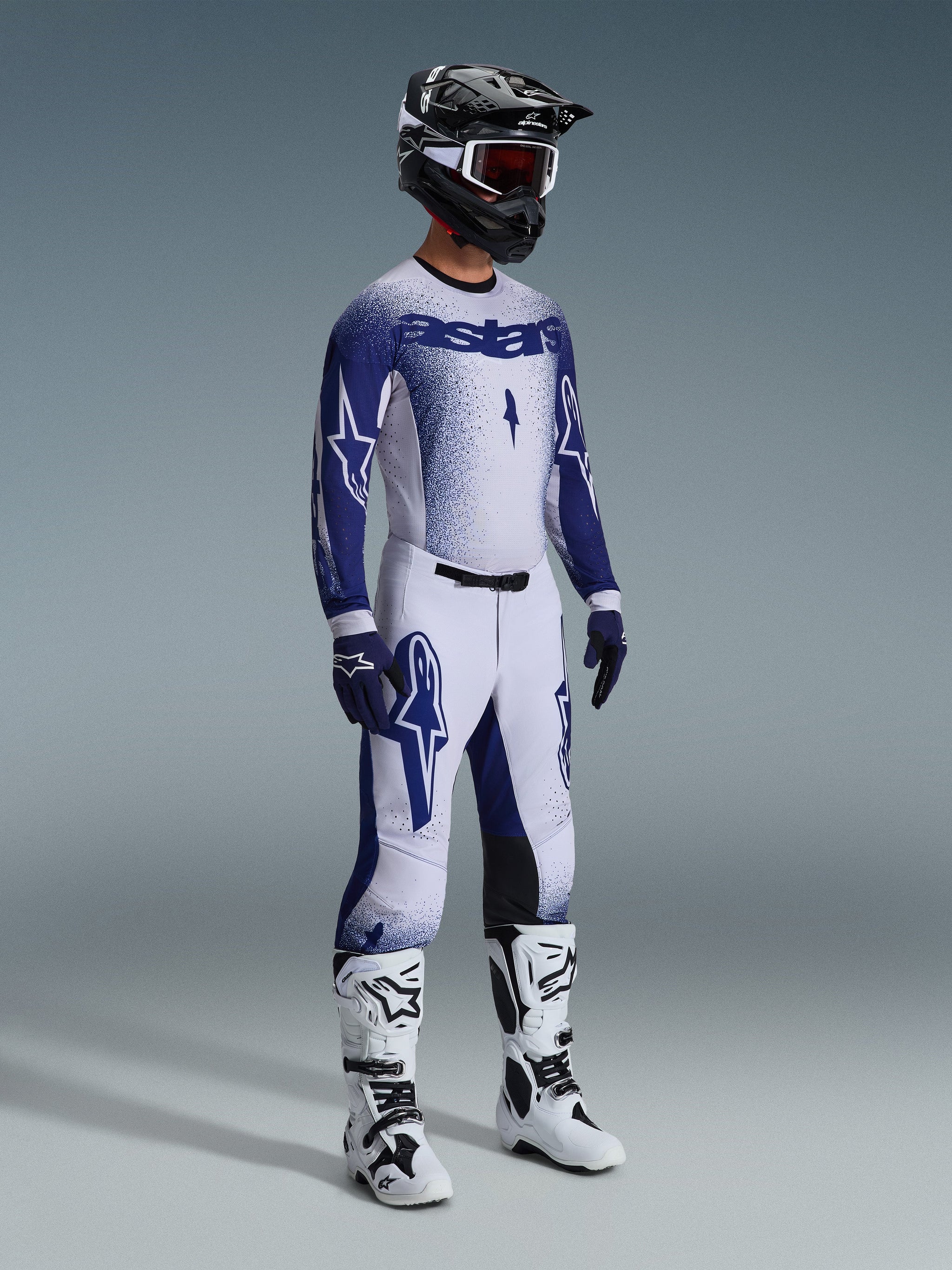 Alpinestars motorcrosser in complete professionele race-uitrusting, 2026 Supertech Scenz Jersey in lichtgrijs en donkerblauw, bijpassende crossbroek met ster-graphics, zwarte helm met rode crossbril, witte en zwarte performance-laarzen en gecoördineerde blauwe en witte handschoenen voor off-road racing.