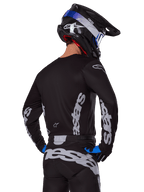 Alpinestars 2025 Racer Graphite Gear Set, motocross-rijder, achteraanzicht, zwart jersey en crossbroek met grijze verticale Alpinestars-logo's, zwarte helm met blauwe en witte accenten en zwarte en blauwe handschoenen, gecoördineerde motorcross uitrusting