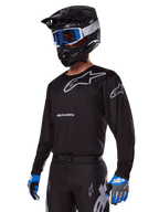 Motorcrosser in Alpinestars 2025 Racer Graphite Gear Set, zwart shirt met grijze Alpinestars-logo's, bijpassende zwarte broek met grijze accenten, zwarte helm met blauwe en witte bril, blauwe en grijze handschoenen, strakke technische outfit voor off-road racen
