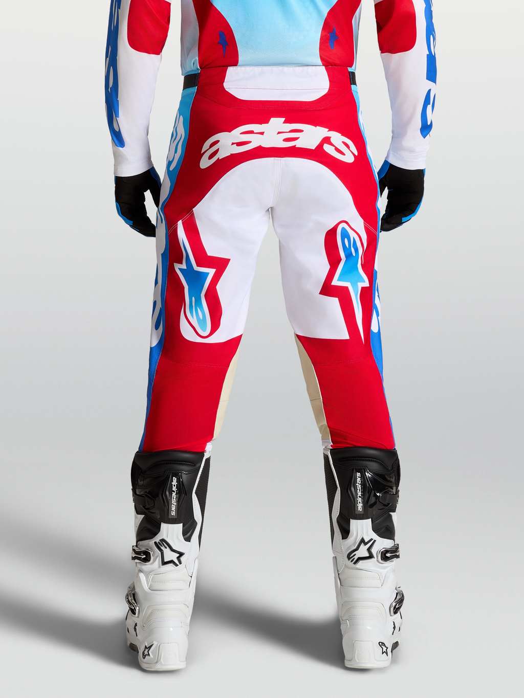 Alpinestars Racer Hunt Broek, rood en wit, voorgevormde motorcrossbroek voor heren met lichtblauwe accenten, achteraanzicht op model met Tech laarzen.