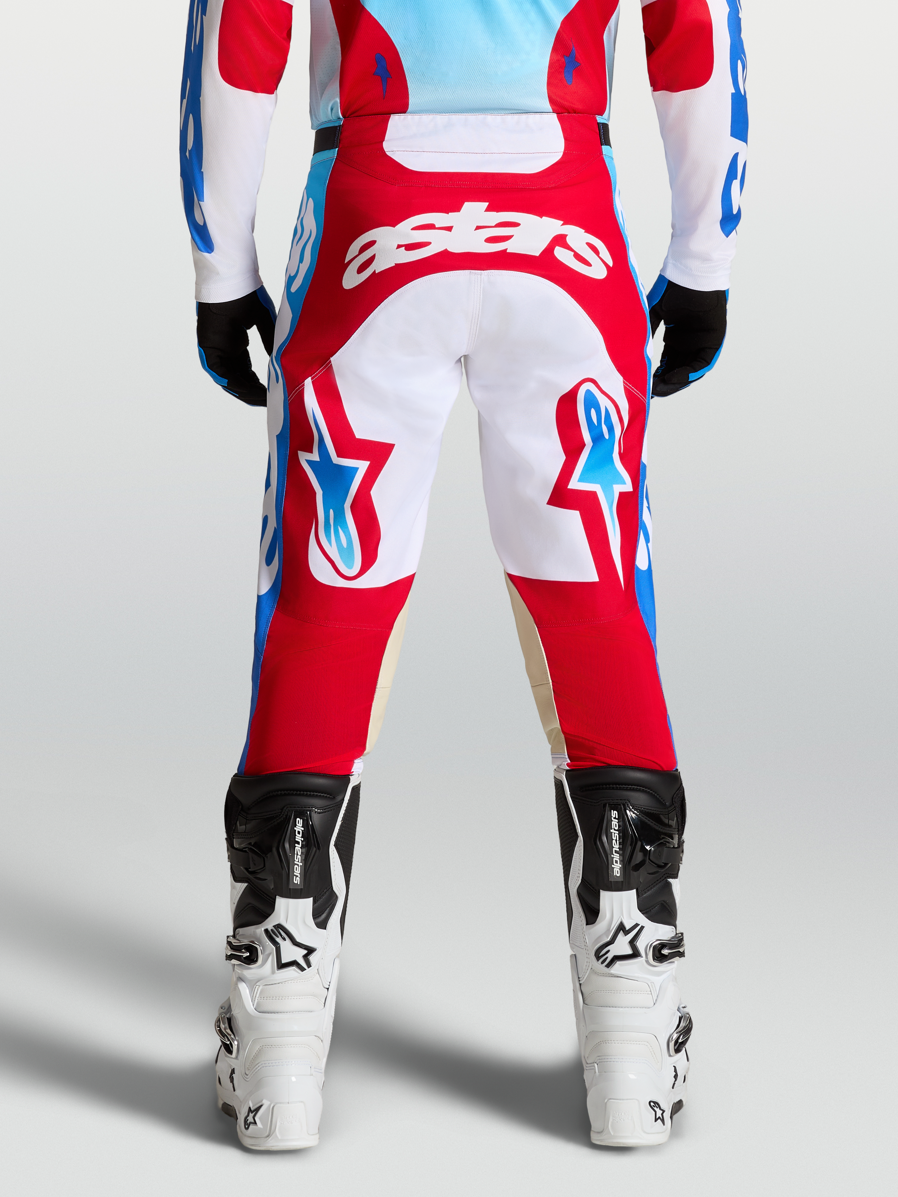 Alpinestars Racer Hunt Broek, rood en wit, voorgevormde motorcrossbroek voor heren met lichtblauwe accenten, achteraanzicht op model met Tech laarzen.