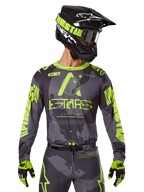 Alpinestars 2025 Racer Hollow Jersey, motorcross raceshirt, donkere camouflage en neongeel, gedragen met bijpassende broek, zwarte en gele helm met crossbril en zwarte handschoenen, voorzien van Alpinestars-branding en camouflage-ontwerp