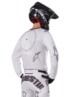 Motocrossrijder vanaf de achterkant in de Alpinestars 2025 Racer Hollow Gear Set in het wit, witte jersey met lange mouwen met grijze sterlogo's en cirkelvormige graphics, bijpassende witte broek met opvallende merknaam op het zitvlak, zwarte helm, gecoördineerde off-road race-uitrusting.