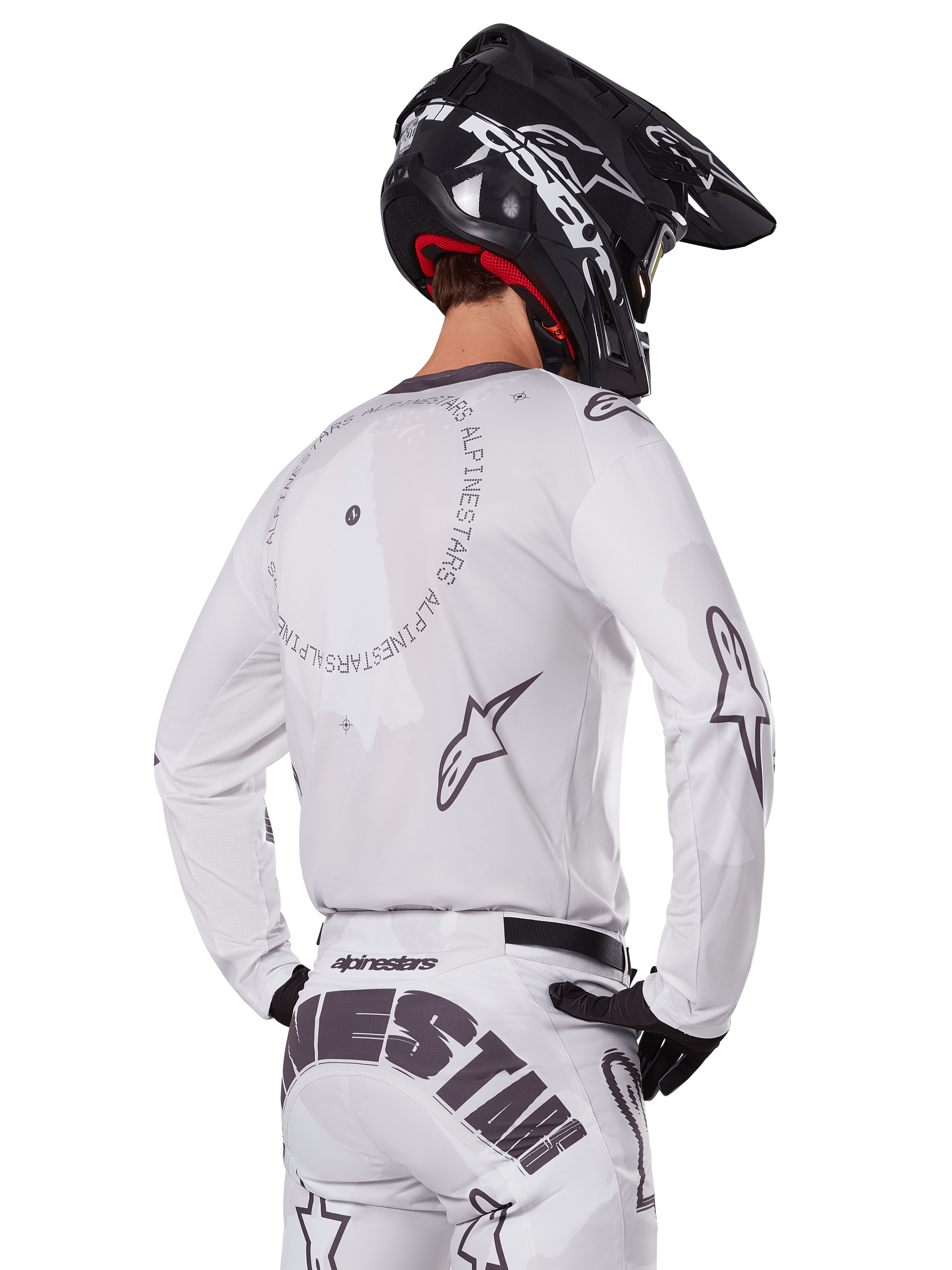 Motocrossrijder vanaf de achterkant in de Alpinestars 2025 Racer Hollow Gear Set in het wit, witte jersey met lange mouwen met grijze sterlogo's en cirkelvormige graphics, bijpassende witte broek met opvallende merknaam op het zitvlak, zwarte helm, gecoördineerde off-road race-uitrusting.