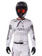 Alpinestars 2025 Racer Hollow Jersey, motocross jersey, wit en zwart, gedragen met een bijpassende witte MX-broek en een zwarte helm met oranje crossbril, voorzien van opvallende Alpinestars-branding en wereldbol-graphics, professionele off-road race-uitrusting