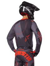 Alpinestars 2025 Racer Hollow Jersey, achteraanzicht van een motorcrosser in Gray Camo en Orange Fluo uitrusting, voorzien van een bijpassende camouflagebroek met grote oranje Alpinestars-letters en een zwarte helm, professioneel off-road kledingontwerp