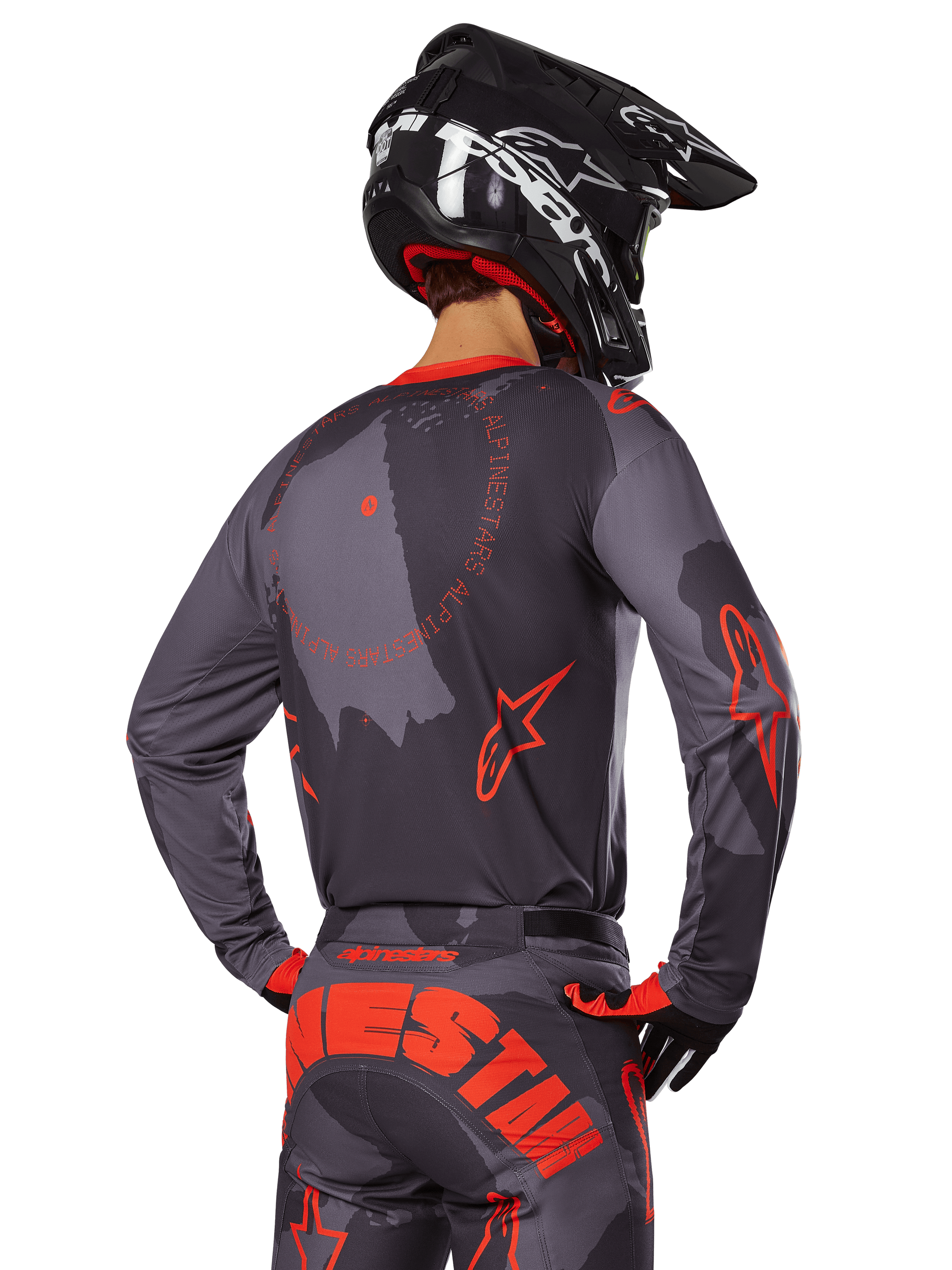 Alpinestars 2025 Racer Hollow Jersey, achteraanzicht van een motorcrosser in Gray Camo en Orange Fluo uitrusting, voorzien van een bijpassende camouflagebroek met grote oranje Alpinestars-letters en een zwarte helm, professioneel off-road kledingontwerp