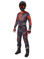 Motorcrossrijder in volledige race-uitrusting, Alpinestars 2025 Racer Hollow Jersey en broek in grijs camo en fluo oranje, zwarte helm met oranje bril, zwart-rode handschoenen en zwarte laarzen met witte logo's, gecoördineerde prestatie-outfit voor off-road racing