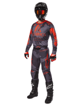 Motorcrossrijder in volledige race-uitrusting, Alpinestars 2025 Racer Hollow Jersey en broek in grijs camo en fluo oranje, zwarte helm met oranje bril, zwart-rode handschoenen en zwarte laarzen met witte logo's, gecoördineerde prestatie-outfit voor off-road racing