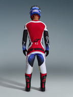 Motocross rider wearing the Alpinestars 2026 Techstar Knif Gear Set, viewed from behind, featuring a red white and black jersey with blue gradient accents on the sleeves, matching white and black pants with bold red branding, blue helmet, and black and red boots, professional off-road racing outfit.