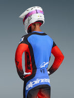 Motocross rider seen from behind wearing the 2026 Techstar Knif Gear Set, blue jersey with red sleeves and black accents, black and blue pants with white Alpinestars branding, white helmet with pink goggle strap, featuring laser-cut perforations for ventilation