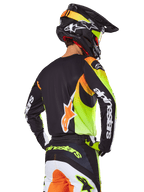 Alpinestars 2025 Fluid Wurx Gear Set, motorcrossrijder van achteren gezien, zwarte jersey met felle neongele en oranje graphics, bijpassende broek met witte en oranje panelen, zwarte helm met neonaccenten, opvallende Alpinestars branding op de mouwen en het zitvlak