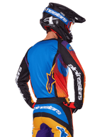 Alpinestars 2025 Fluid Wurx Gear Set, motorcrossrijder van achteren gezien, meerkleurige jersey met blauwe achterkant en zwarte mouwen met witte logo's, bijpassende oranje en blauwe broek, zwart-blauwe helm met oranje bril, gecoördineerde off-road race-outfit