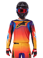 Alpinestars 2025 Fluid Wurx Jersey, multicolor motorcross jersey met oranje en rood kleurverloop en paarse accenten, gedragen met bijpassende uitrusting, zwarte helm met blauwe en rode bril en rode handschoenen, groot zwart sterlogo op de borst