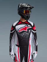 Alpinestars 2026 Techstar Nomur Jersey, motorcross shirt, zwart wit en rood, gedragen door een rijder met bijpassende broek, zwarte helm en witte handschoenen, voorzien van een groot rood Astars-logo en gespikkeld grafisch ontwerp, atletische racepasvorm voor off-road competitie