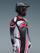 Motorcrosser in de Alpinestars 2026 Techstar Nomur Jersey in zwart, wit en rood, voorzien van een groot rood Astars-logo en gespikkelde grafische patronen, gecombineerd met een bijpassende crossbroek, witte handschoenen en een zwarte helm met bril, gecoördineerde outfit voor off-road competitie.