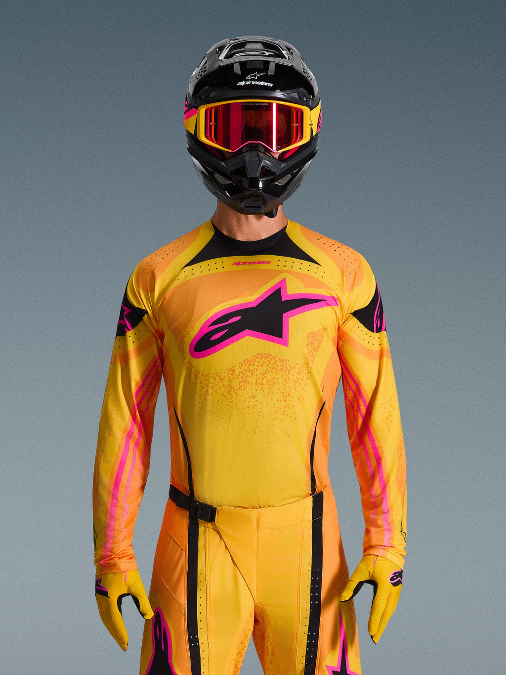 Alpinestars 2026 Techstar Nomur Gear Set, Motocross rider wearing yellow and orange jersey and pants with bright pink accents, black helmet with yellow and pink graphics, red-tinted goggles with yellow frames, coordinated performance racing outfit