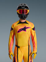 Alpinestars 2026 Techstar Nomur Jersey in oranje, geel en roze, gedragen door een motorcrosser met een bijpassende zwarte helm en roze bril, voorzien van een groot roze Alpinestars-logo op de borst, atletische racepasvorm voor off-road prestaties.