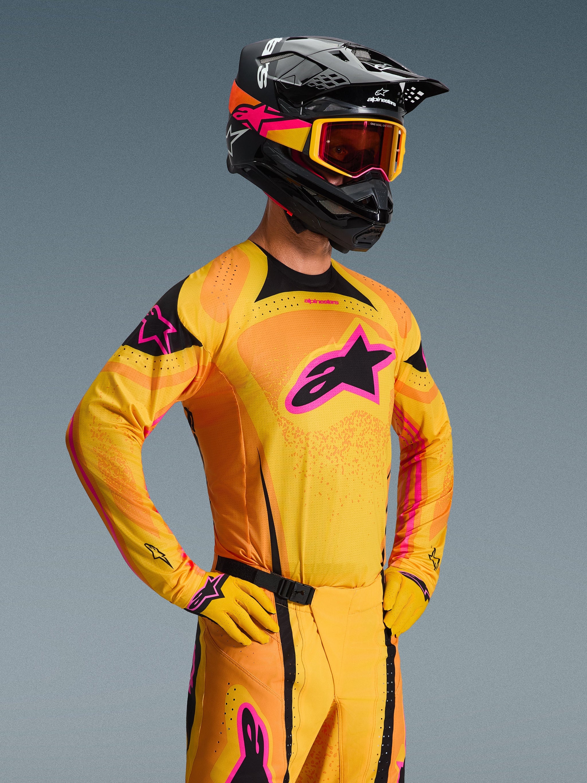 Alpinestars 2026 Techstar Nomur Jersey, motorcross shirt, oranje, geel en roze, gedragen door een rijder met een bijpassende zwarte helm en crossbril, voorzien van een groot roze sterlogo op de borst, zwarte accenten en lasergesneden ventilatiedetails.