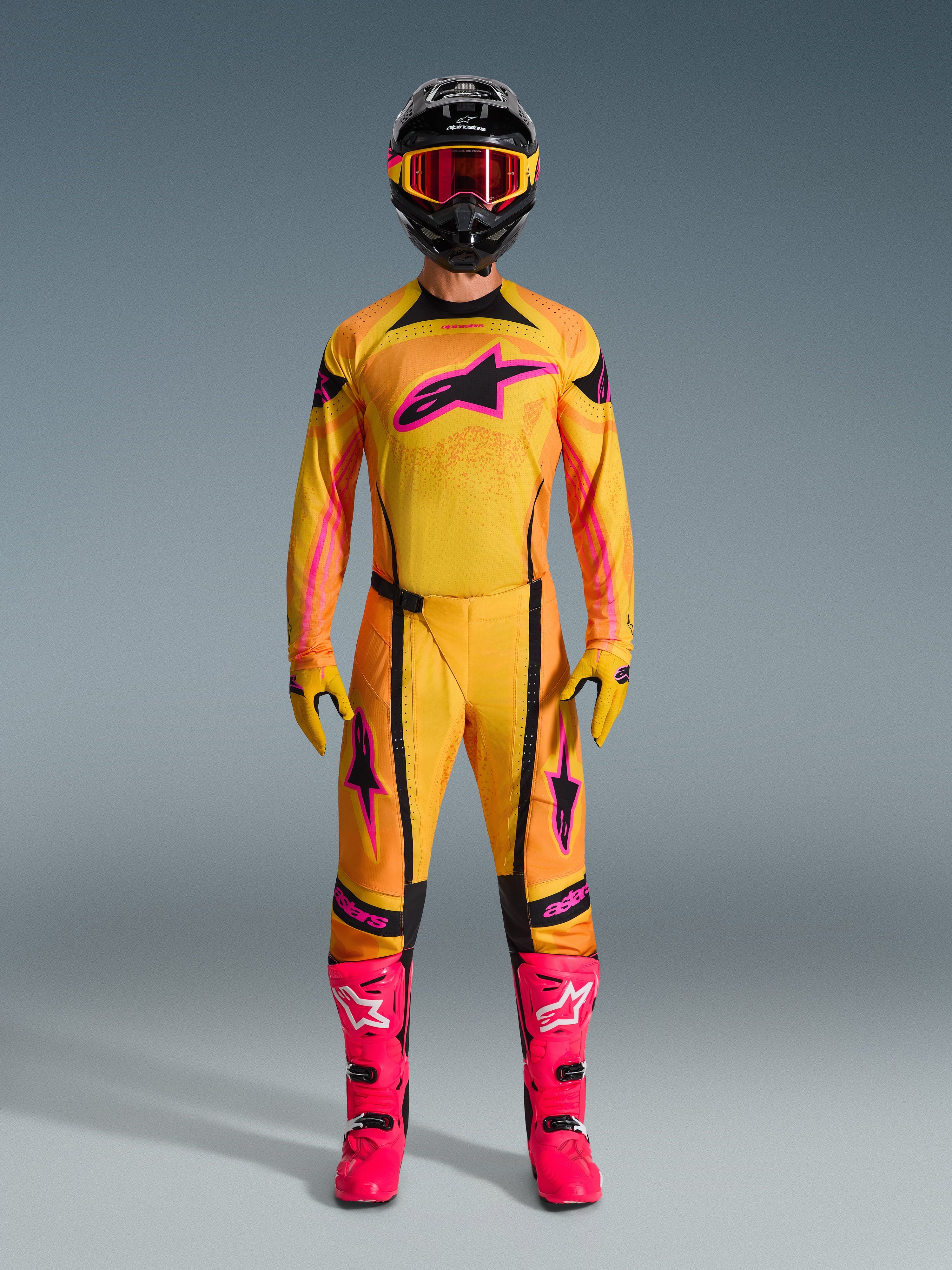 2026 Techstar Nomur Gear Set, complete motocross racing apparel worn by a rider, yellow and orange gradient jersey with pink star logo, matching pants and gloves, bright pink boots and black helmet, professional MX kit on a gray background