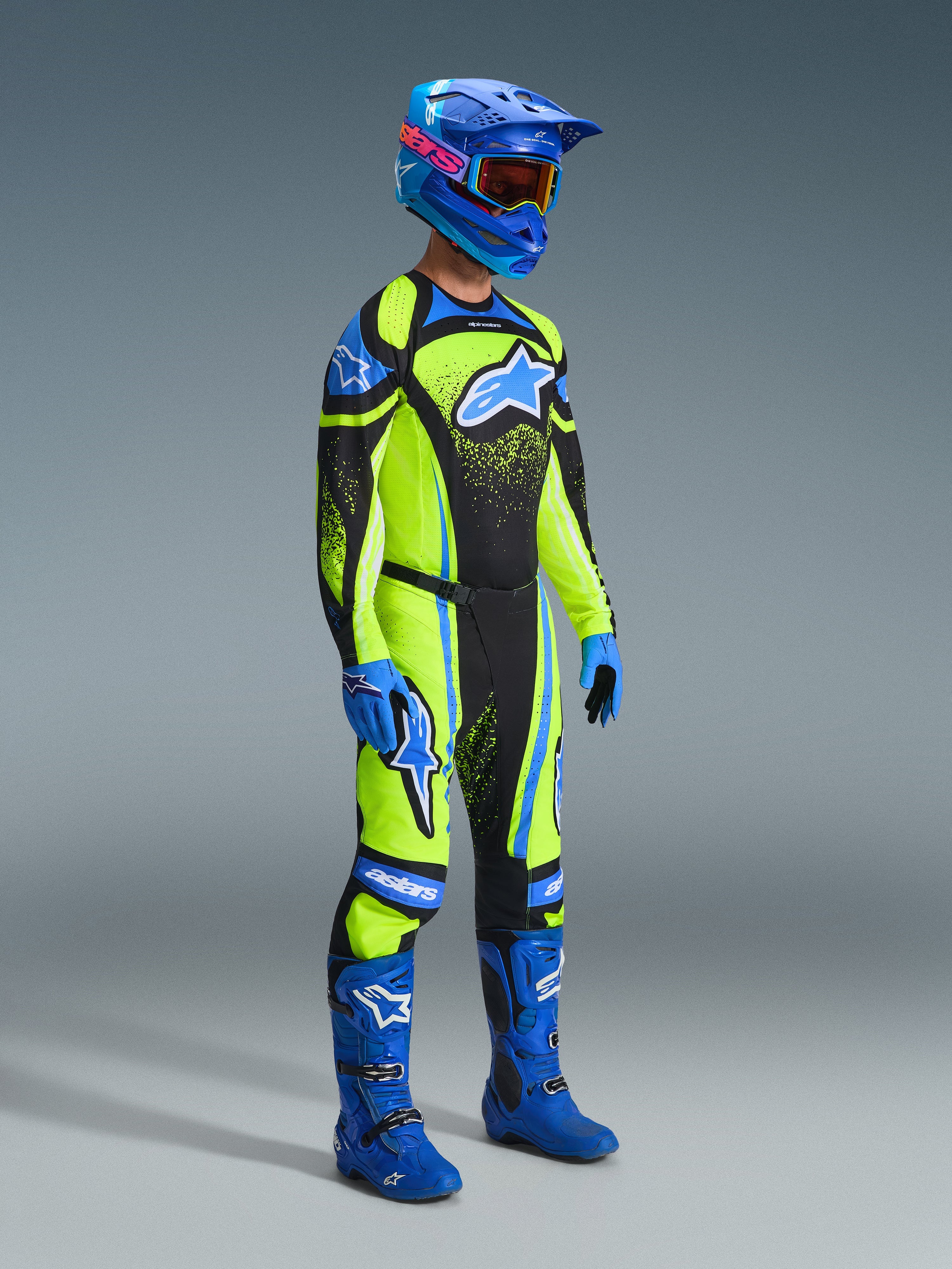 Motocross rider wearing the Alpinestars 2026 Techstar Nomur Gear Set, bright lime green jersey and pants with black panels and blue accents, large blue star logo on chest and thighs, blue helmet with orange goggles, blue boots, and blue gloves, full racing kit for motocross