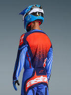 Motocross rider from behind wearing the 2026 Techstar Nomur Jersey, Dark Navy with Orange and UCLA Blue accents, featuring a large orange textured graphic on the back, paired with matching motocross pants and a blue racing helmet, coordinated professional gear for off-road riding.