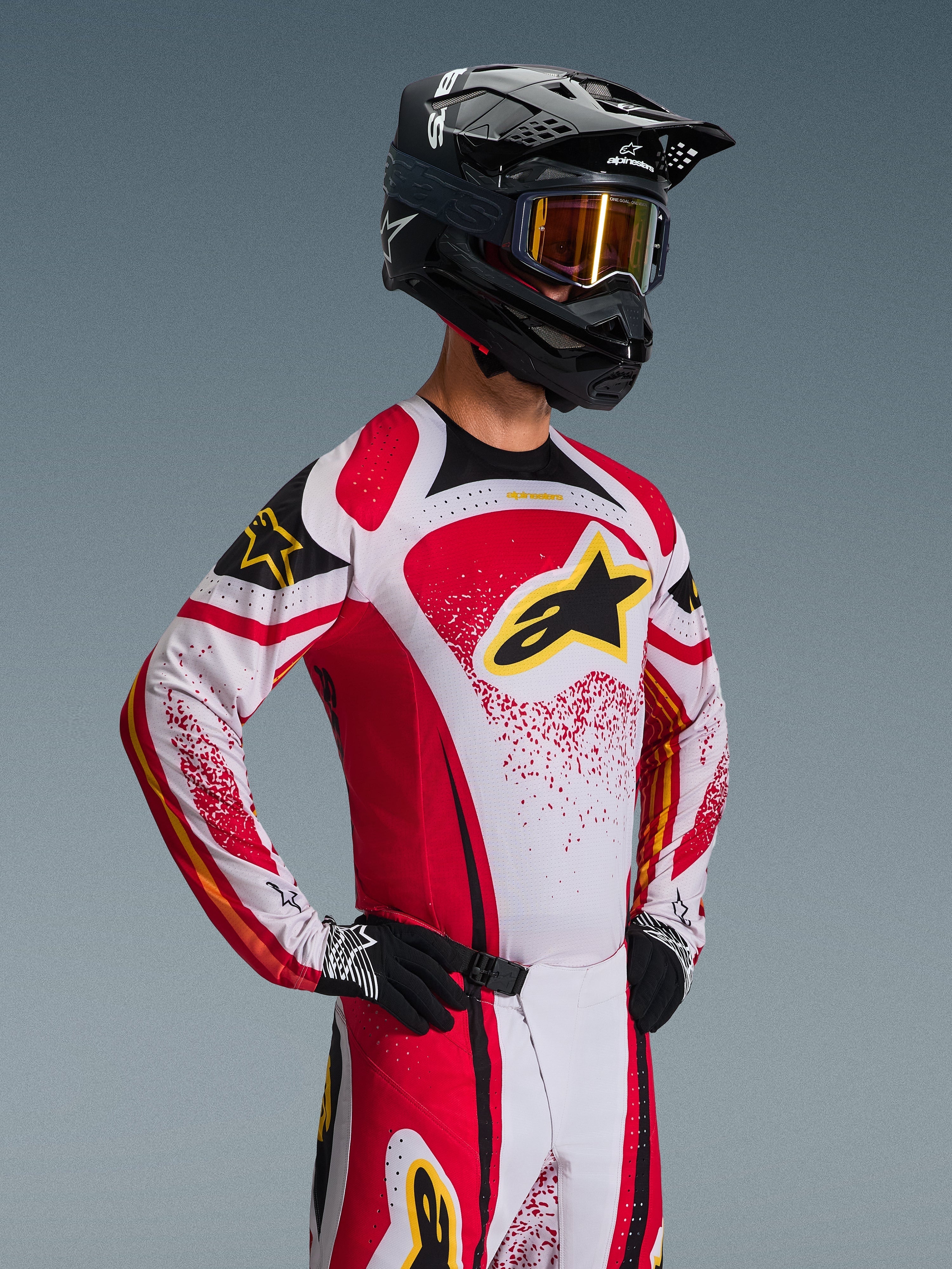 Motocross rider wearing the Alpinestars 2026 Techstar Nomur Jersey, Red White and Black with yellow accents, paired with matching pants, a black helmet with gold goggles, and black gloves, athletic race fit for professional off-road competition