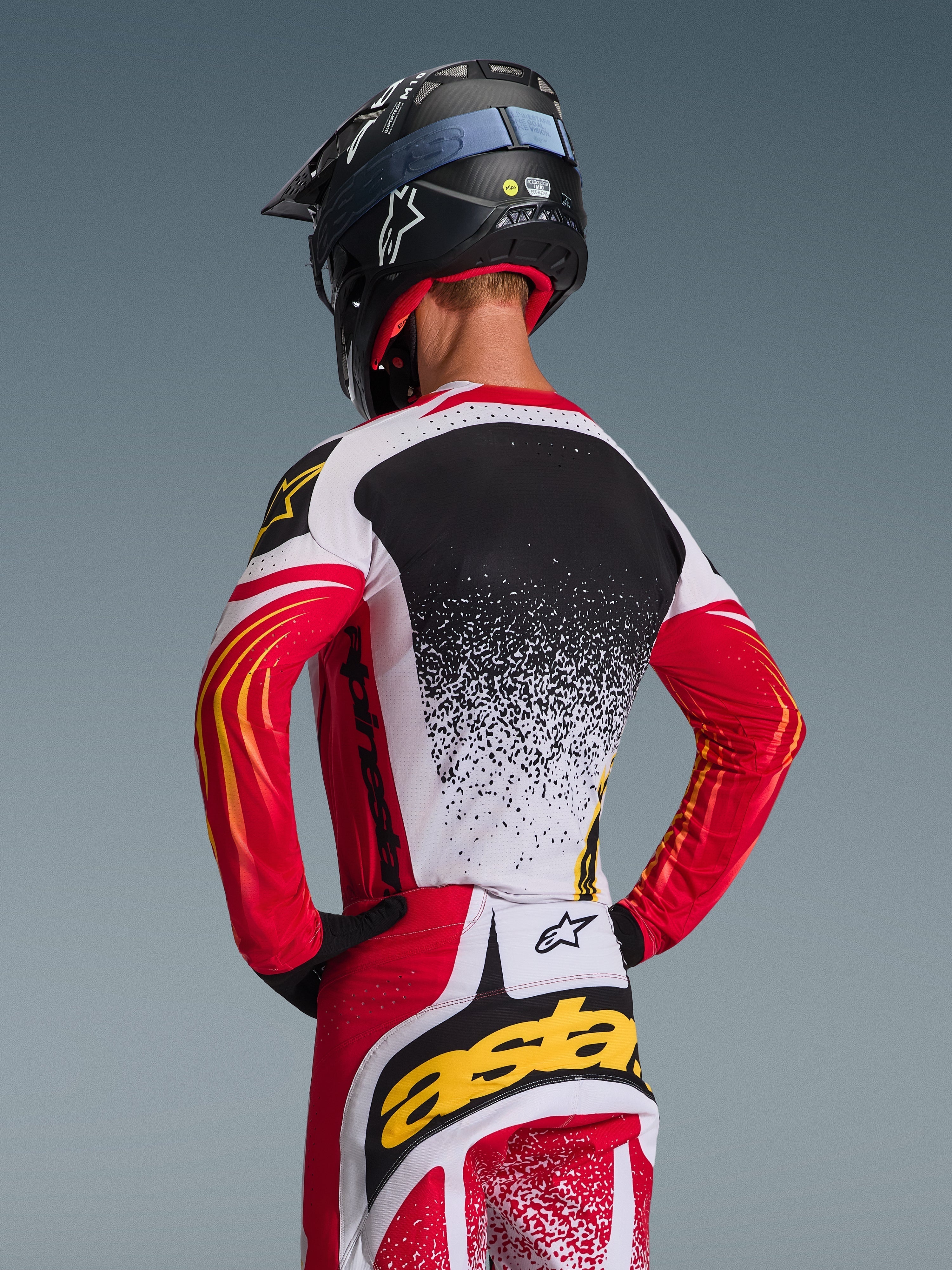 Motocross rider from behind wearing the Alpinestars 2026 Techstar Nomur Jersey and matching pants, Red White and Black with yellow accents, featuring a speckled gradient design on the back, black Alpinestars helmet and black gloves, coordinated professional gear for off-road racing