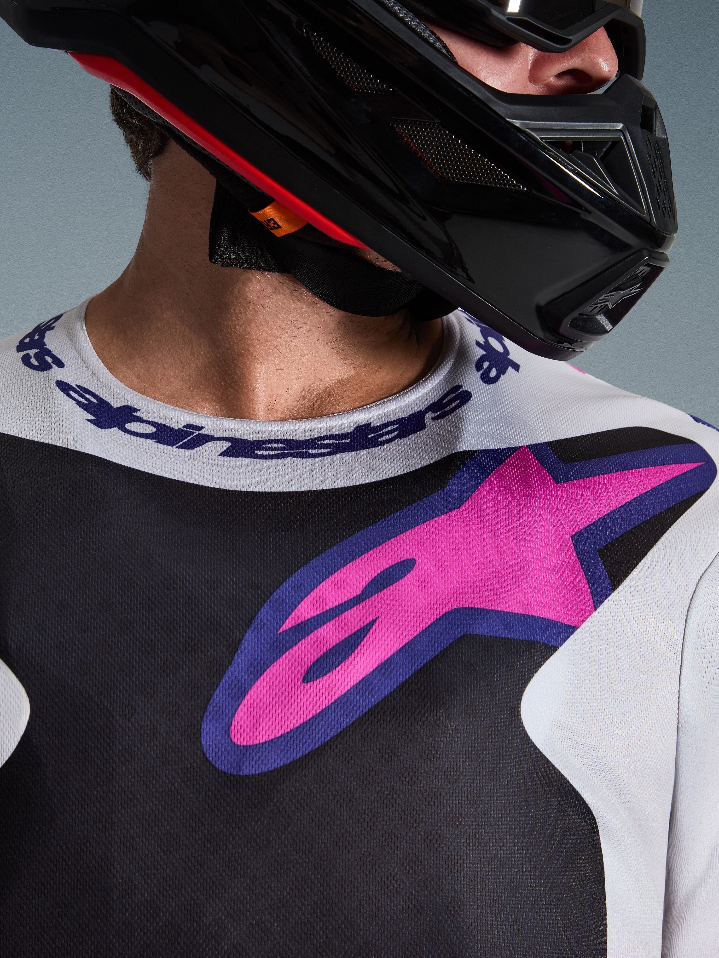 Close-up of Alpinestars 2026 Fluid Grid Jersey, Motocross Jersey, Black Light Gray and Purple, featuring a vibrant pink-purple star logo on the chest and branded sport collar, worn with a black motocross helmet