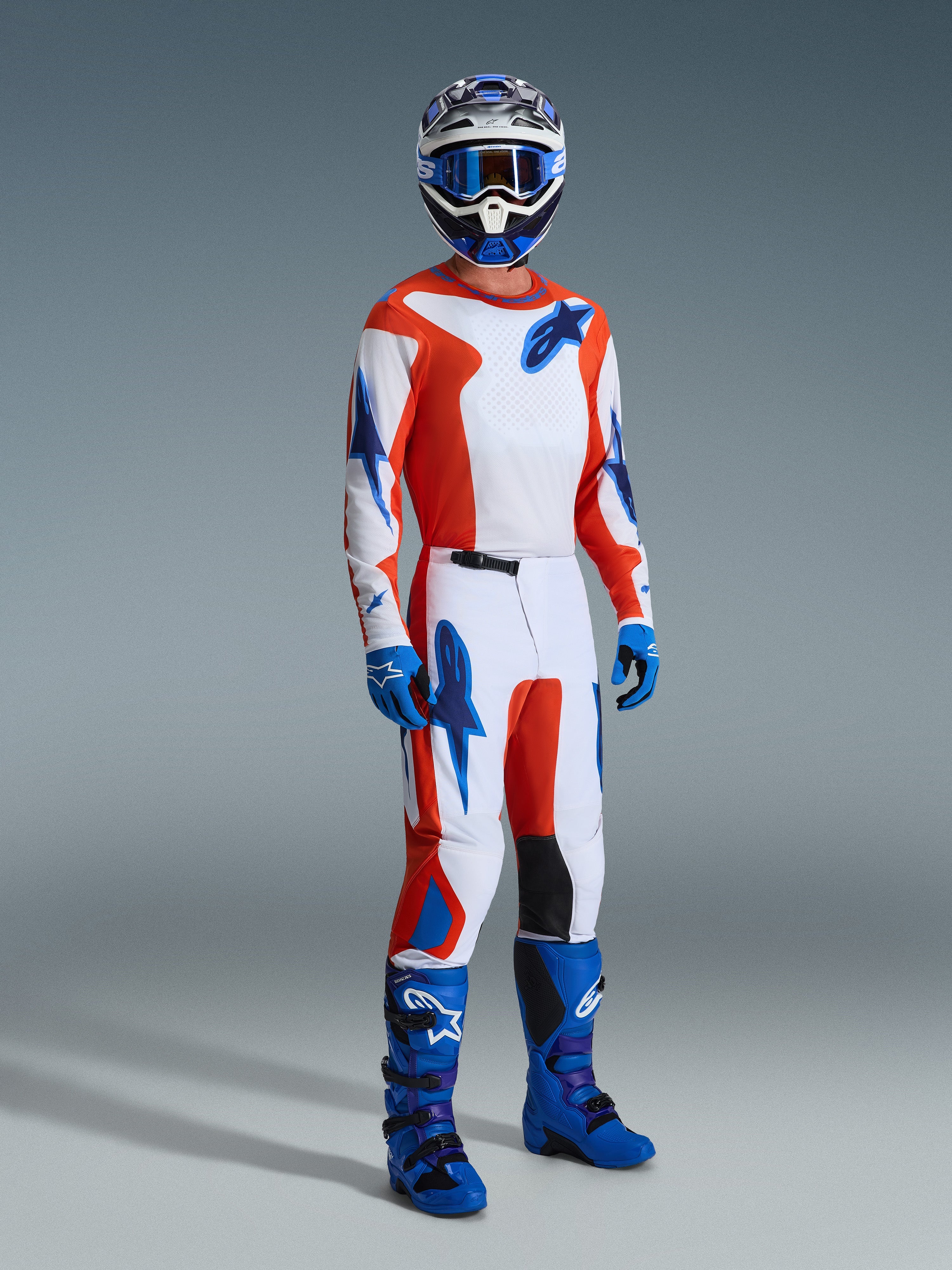 Motocross rider wearing the Alpinestars 2026 Fluid Grid Gear Set, white jersey and pants with orange and UCLA blue graphics, matching blue helmet and goggles, blue gloves, and bright blue racing boots, coordinated performance outfit for off-road riding.
