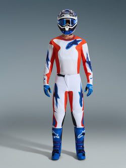 Alpinestars 2026 Fluid Grid crossuitset, motorcrossrijder in volledige uitrusting, witte jersey en broek met oranje en UCLA-blauwe accenten, bijpassende blauw-witte helm met blauwe crossbril, blauwe handschoenen en blauwe motorcrosslaarzen, gecoördineerde off-road race-outfit