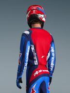 Rear view of a motocross rider wearing the Alpinestars 2026 Fluid Grid Jersey in Blue and Red, featuring bold white branding on the sleeves and back, paired with matching motocross pants and a red helmet for a complete off-road racing look