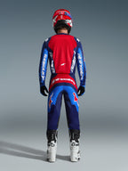 Rear view of a motocross rider wearing the 2026 Fluid Grid Jersey in Blue and Red, featuring a large red back panel and blue sleeves with white Alpinestars branding, paired with matching pants, white and black boots, and a red helmet for a complete off-road racing outfit.
