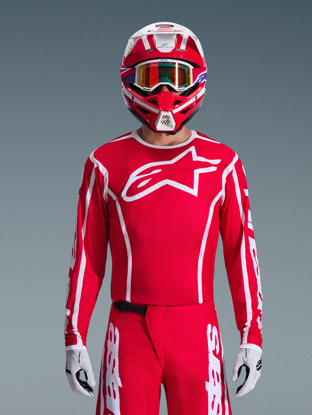 Motocross rider wearing Alpinestars 2026 Fluid Apex Gear Set, Red jersey with large white star logo and white piping, matching red pants with white accents, red and white helmet with purple highlights and orange-tinted goggles, white and black gloves, coordinated outfit for off-road racing