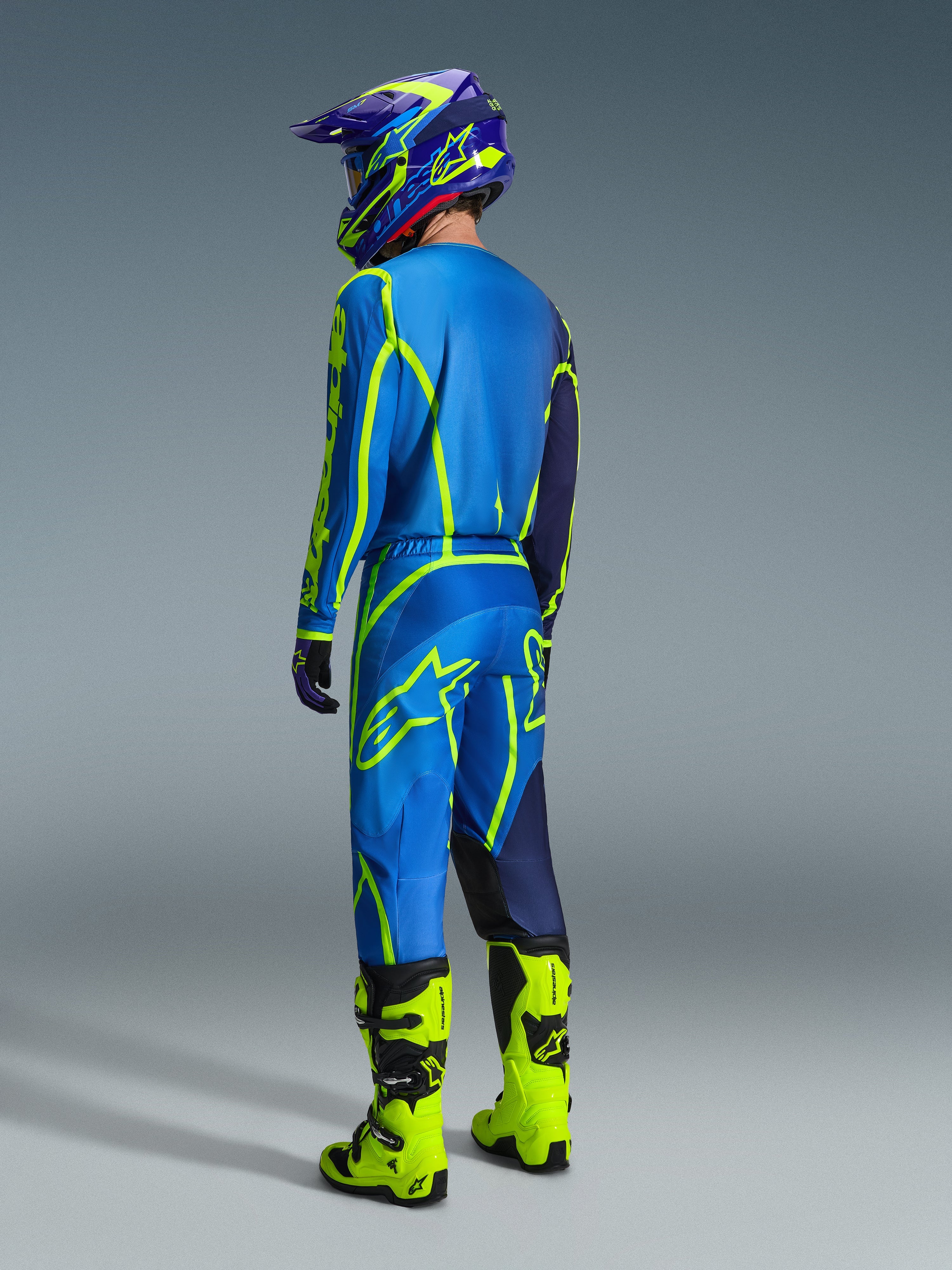 Alpinestars 2026 Fluid Apex Gear Set, motocross rider shown from the back wearing a blue and neon yellow gear set, blue jersey and pants with neon yellow logos, neon yellow boots, purple and blue helmet, coordinated off-road racing apparel.