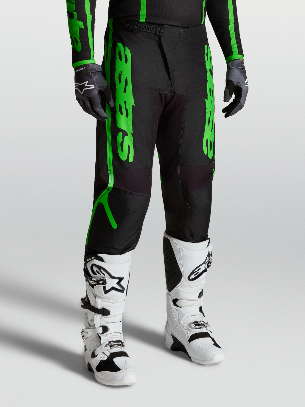 Alpinestars Fluid Apex V2 Broek, motocrossbroek voor heren, zwart en neon groen, gedragen met witte motorlaarzen en zwarte handschoenen, duurzame 600D polyfabric constructie.