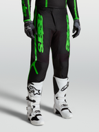 Alpinestars Fluid Apex V2 Broek, motocrossbroek voor heren, zwart en neon groen, gedragen met witte motorlaarzen en zwarte handschoenen, duurzame 600D polyfabric constructie.