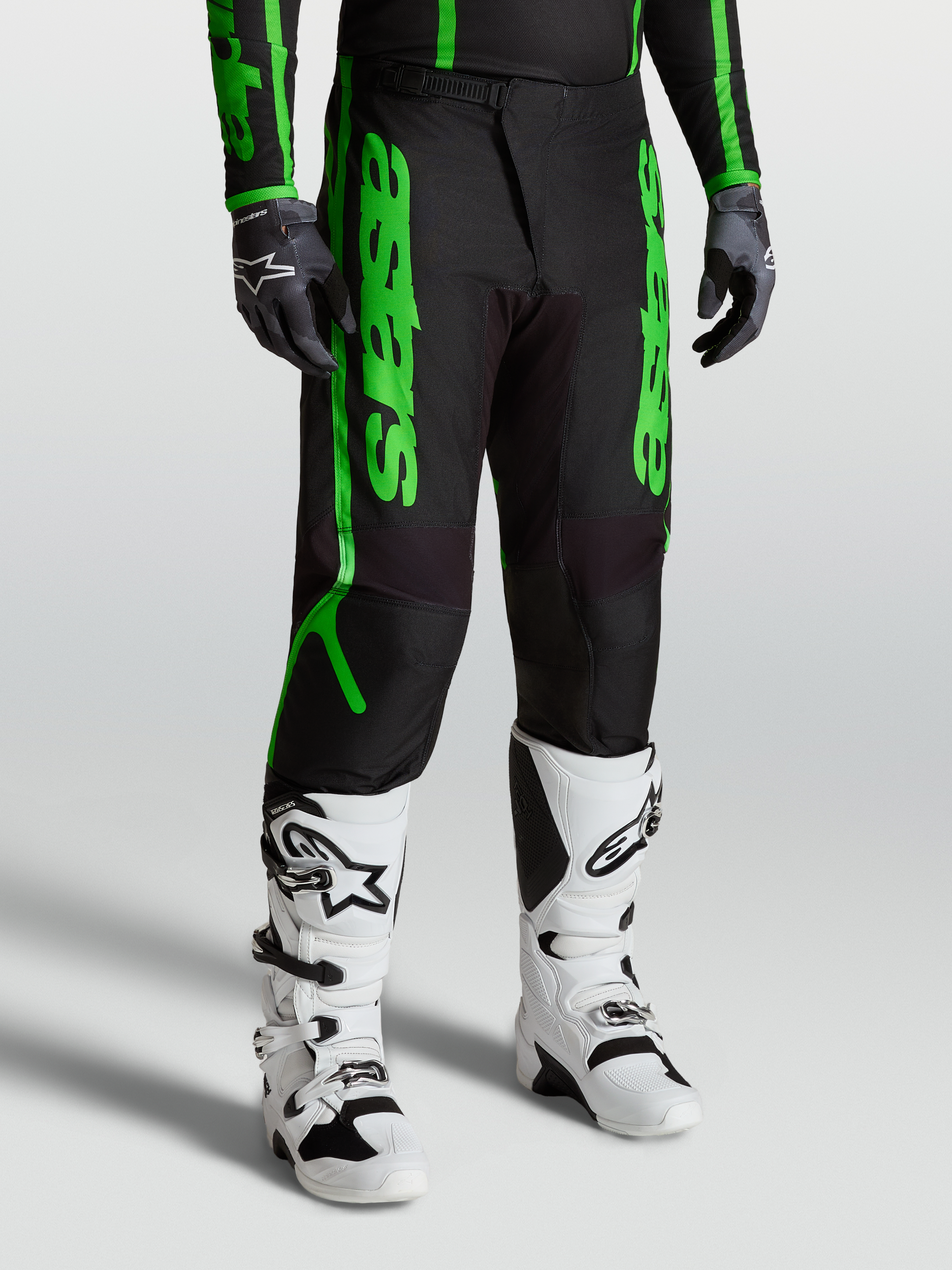 Alpinestars Fluid Apex V2 Broek, motocrossbroek voor heren, zwart en neon groen, gedragen met witte motorlaarzen en zwarte handschoenen, duurzame 600D polyfabric constructie.