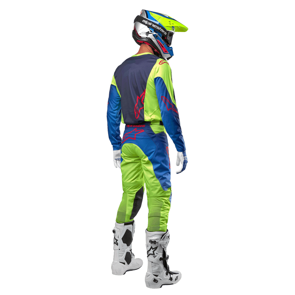 Motocrossrijder van achteren gezien die de Alpinestars 2024 Racer Hoen Jersey draagt in Yellow Fluo en Blue Night Navy, bijpassende motorcrossbroek en witte laarzen, toont het ontwerp aan de achterzijde met rode logo's, gecoördineerde outfit voor professioneel off-road racen