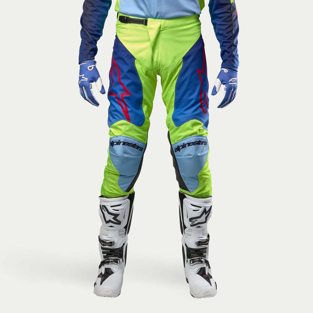 Alpinestars 2024 Racer Hoen broek, motorcrossbroek, fluoriserend geel en nachtblauw, gedragen met witte en zwarte motorcrosslaarzen en blauwe handschoenen, voorzien van opvallende rode logo's, lichtblauwe kniepanelen en een sportief ergonomisch ontwerp voor off-road rijden