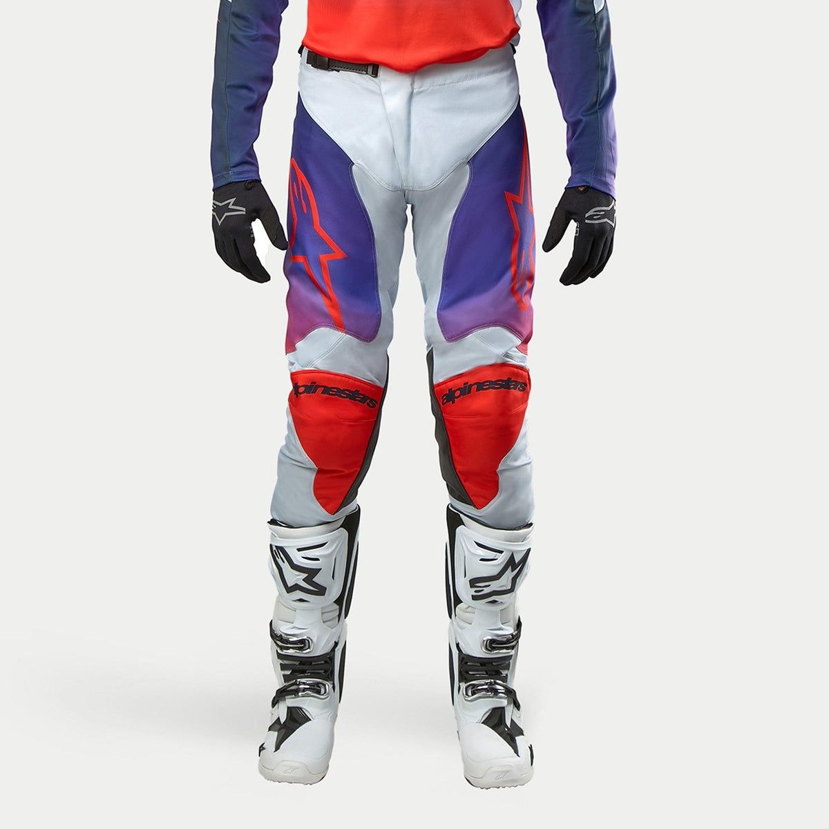 Alpinestars 2024 Racer Hoen Pants, motorcross broek, lichtgrijs met paarse en oranje kleuraccenten, rode kniestukken en zwarte details, persoon draagt complete uitrusting met witte laarzen en zwarte handschoenen, sportieve pasvorm voor off-road rijden