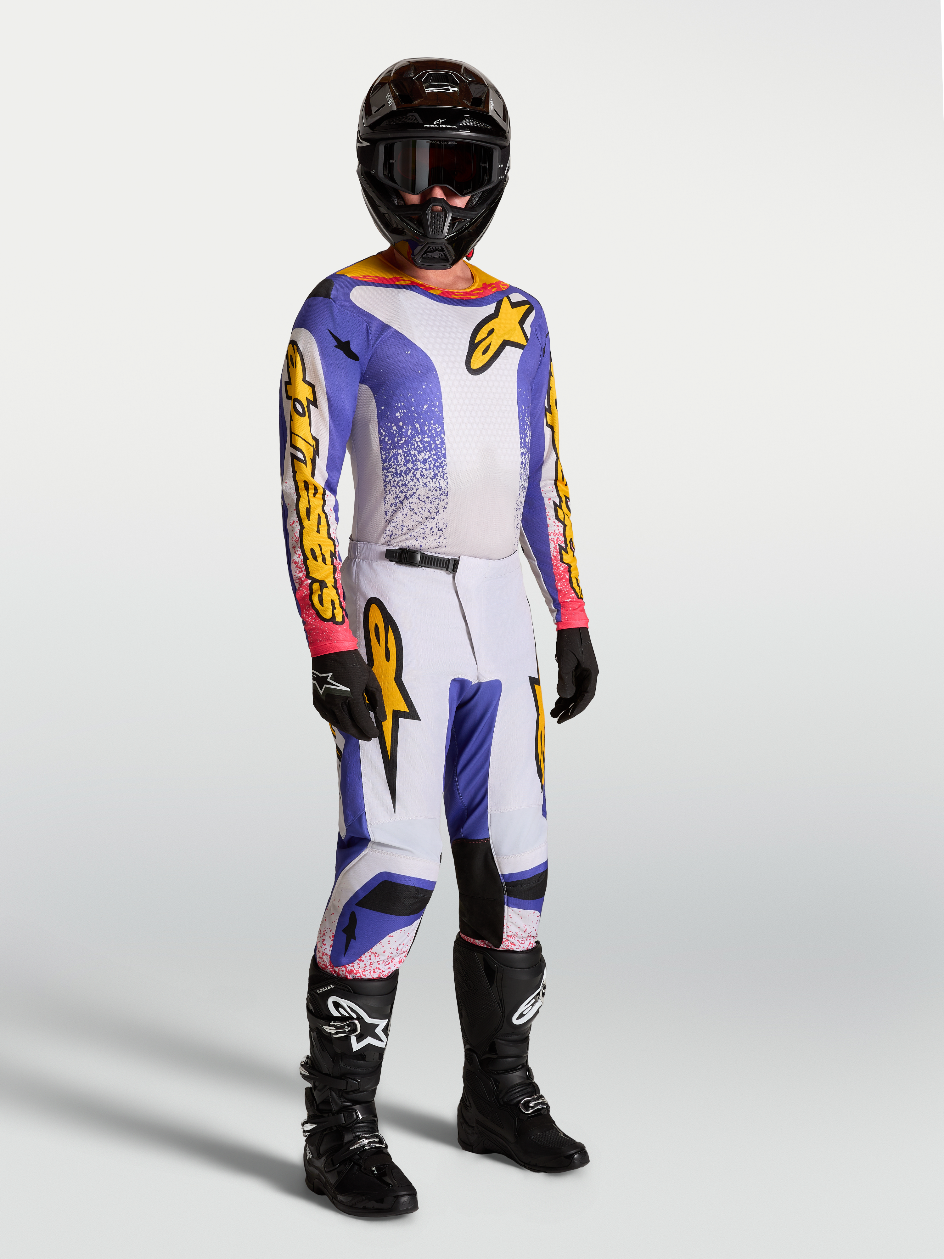 Alpinestars Fluid Drag jersey en broek voor in laarzen, motocross-uitrusting voor heren, licht lila, blauw en wit, met zwarte motorlaarzen en helm.