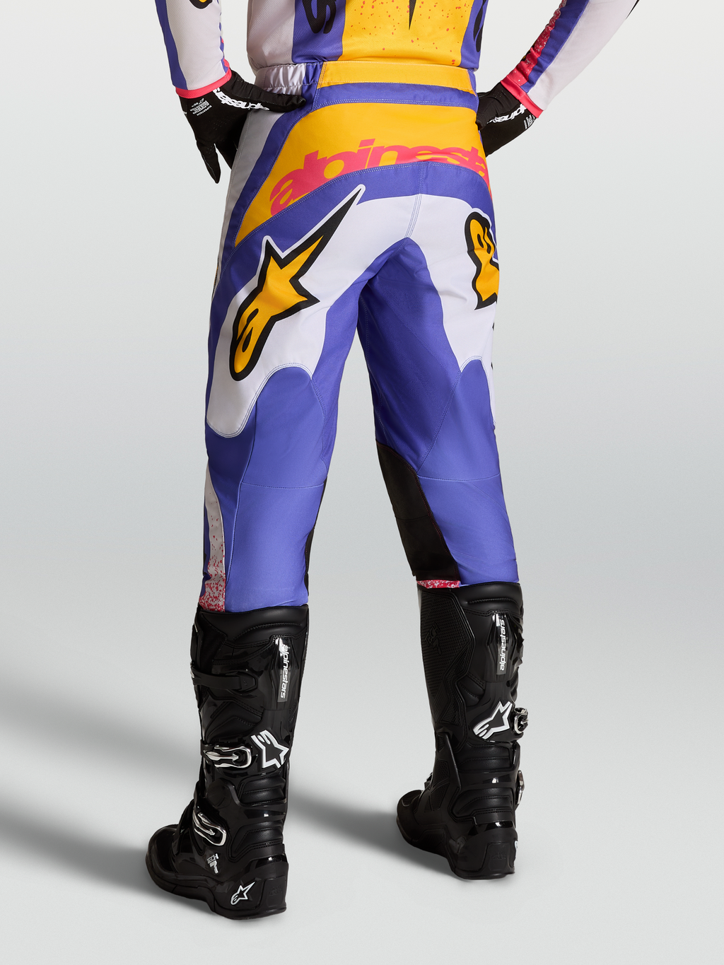 Alpinestars Fluid Drag broek, motorcrossbroek voor heren, blauw en goud met wit, voorzien van versterkt rugpand, gedragen met zwarte Tech 7 laarzen, sportieve pasvorm voor off-road rijden.