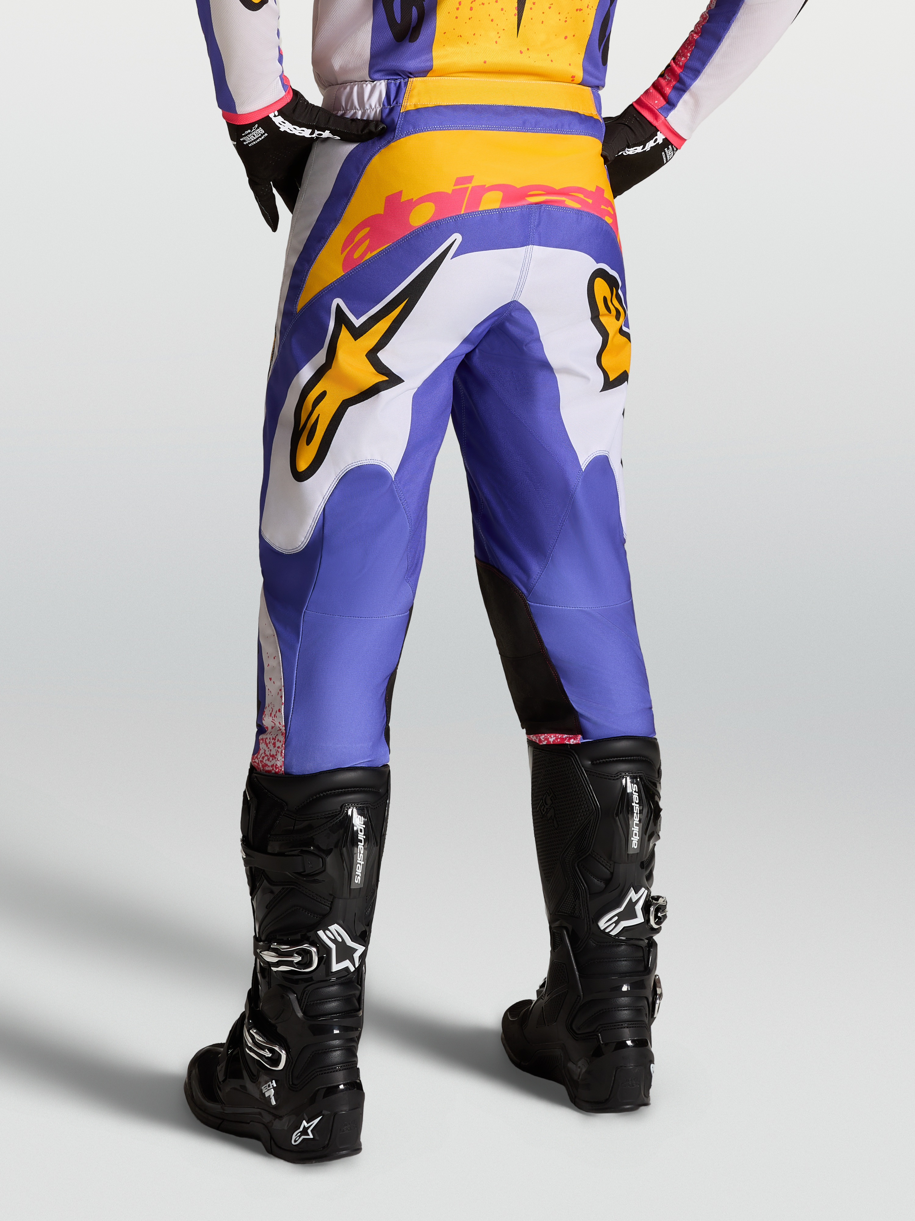 Alpinestars Fluid Drag broek, motorcrossbroek voor heren, blauw en goud met wit, voorzien van versterkt rugpand, gedragen met zwarte Tech 7 laarzen, sportieve pasvorm voor off-road rijden.
