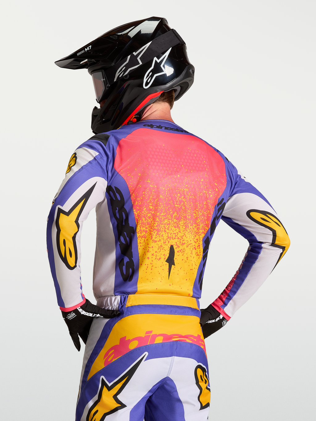 Alpinestars Fluid Drag jersey, blauwe en gouden motorcrossjersey op een model met helm, vochtafvoerend materiaal en ergonomische achterzijde.