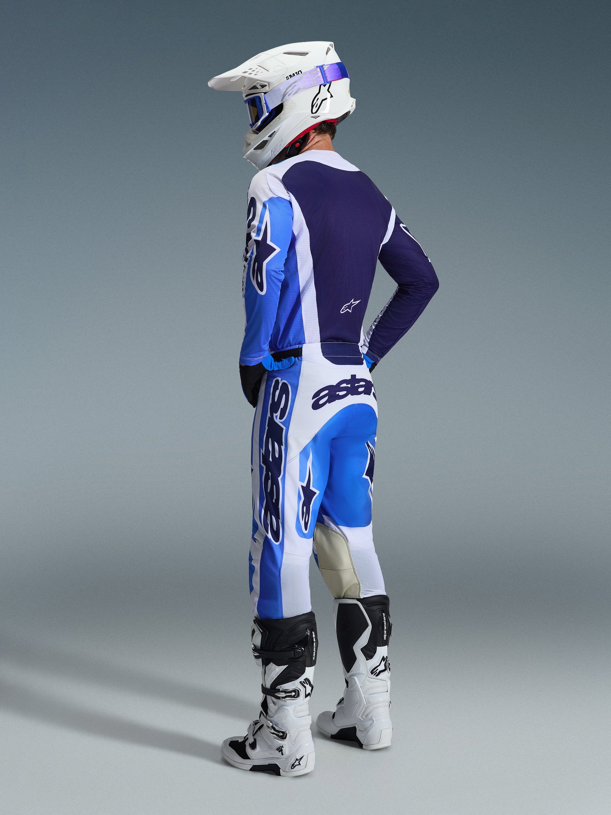 Motocross rider from a rear perspective wearing the Alpinestars 2026 Racer Air Portl Jersey in Ucla Blue and White, paired with matching motocross pants, white Supertech helmet, and white boots, showcasing a complete coordinated racing gear set.